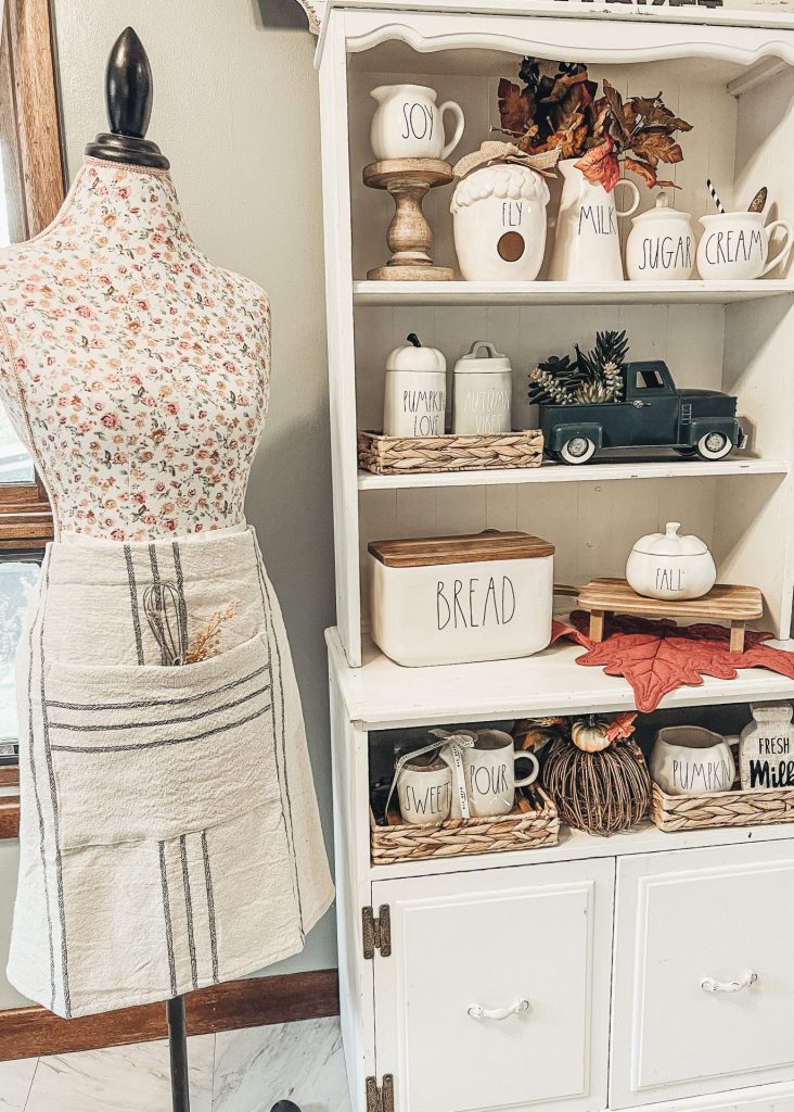 A doll maker’s dummy with a floral pattern next to a small hutch filled with Rae Dunn pottery
